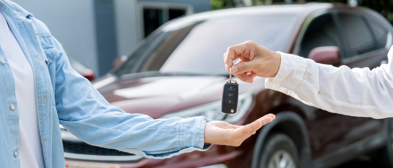 A salesperson giving a key fob to a customer with a burgundy car in the background.