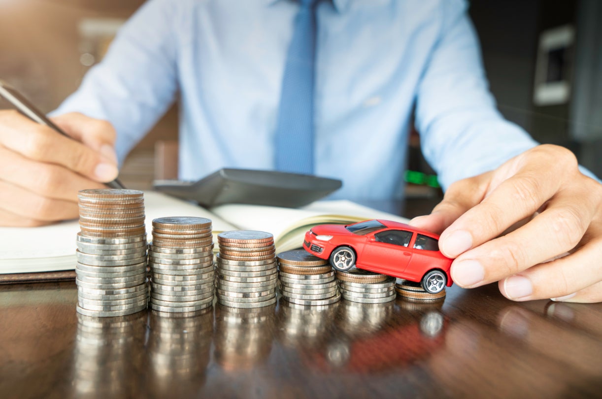 A car dealership salesman driving a red toy car up stacks of coins.